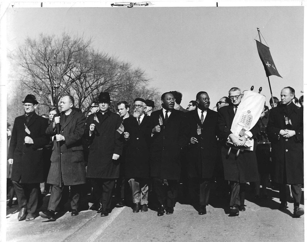 Black and white photo with men marching holding flags and one Torah. Picture includes Martin Luther King, Jr. and Abraham Joshua Heschel.